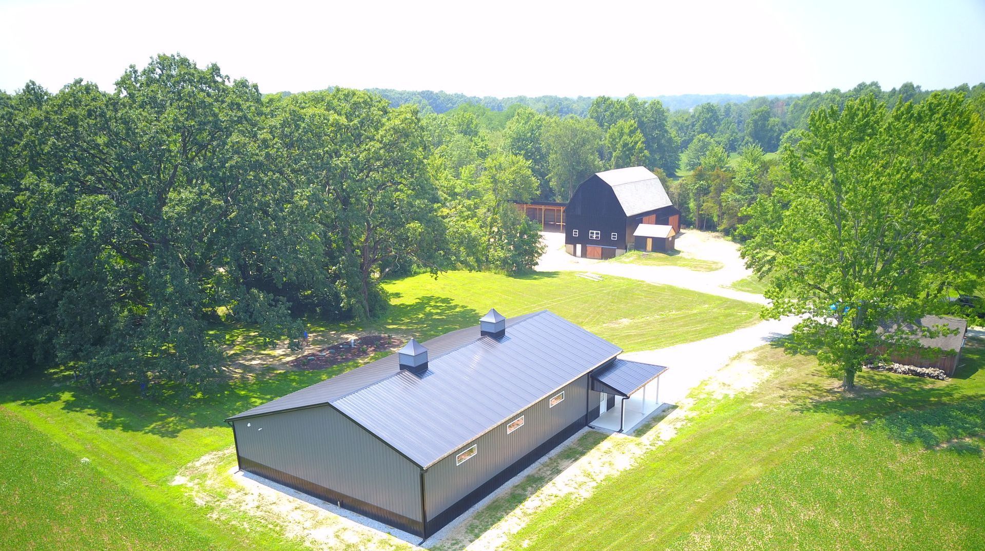 A modern, dark-roofed building sits in a grassy clearing near a wooded area, with a barn in the background.