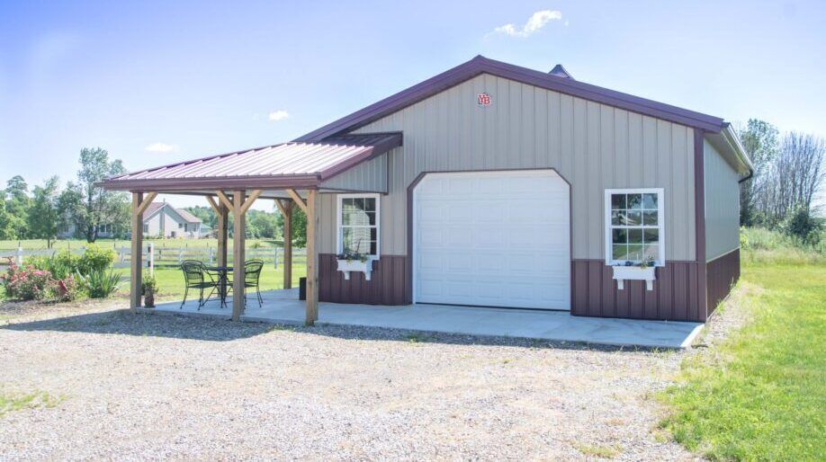 Tan and brown pole barn with a covered porch, a white garage door, and flower boxes, in a grassy area.