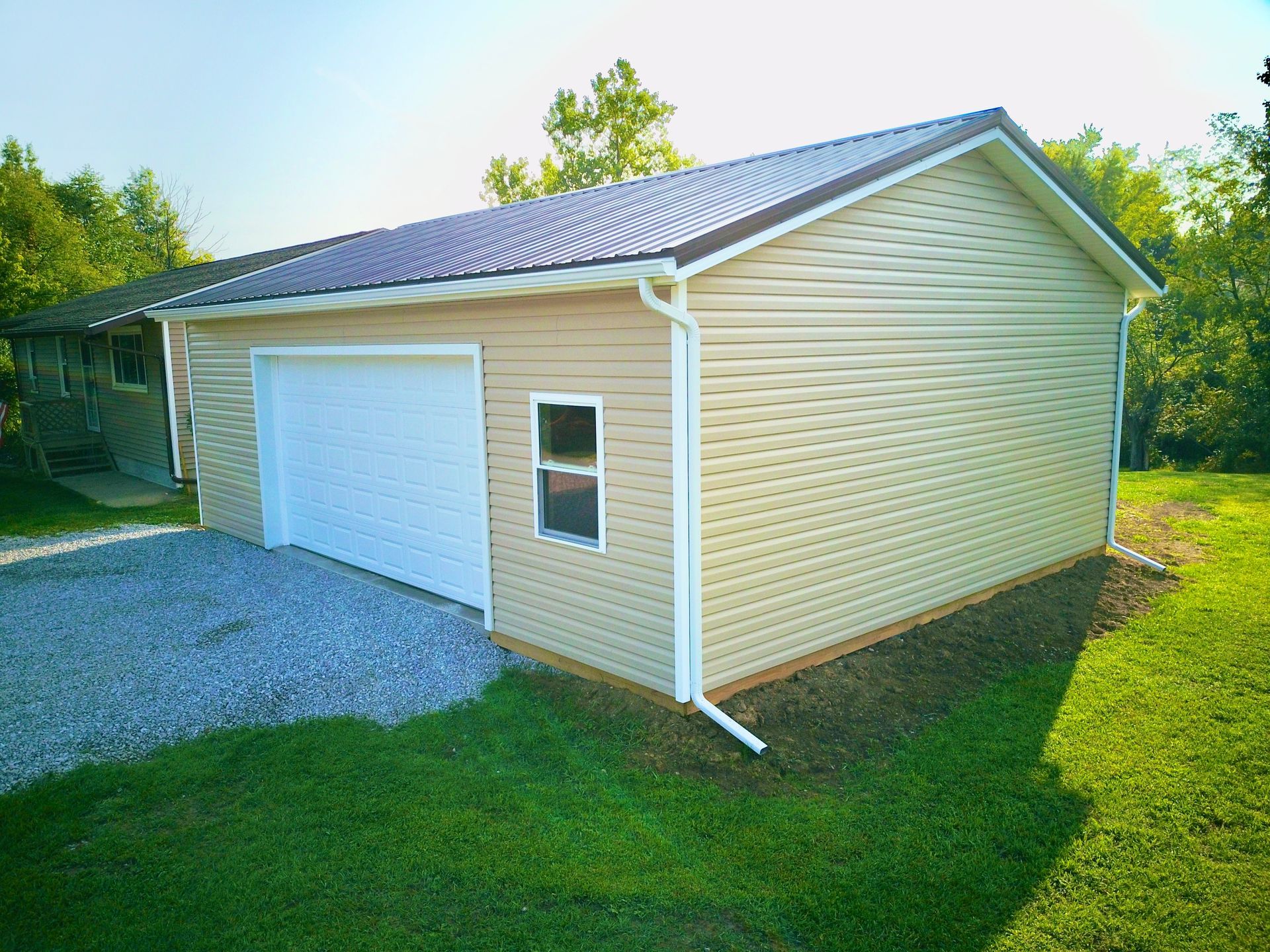 Tan-sided garage with a white garage door and small window. Dark roof, surrounded by grass, gravel driveway.