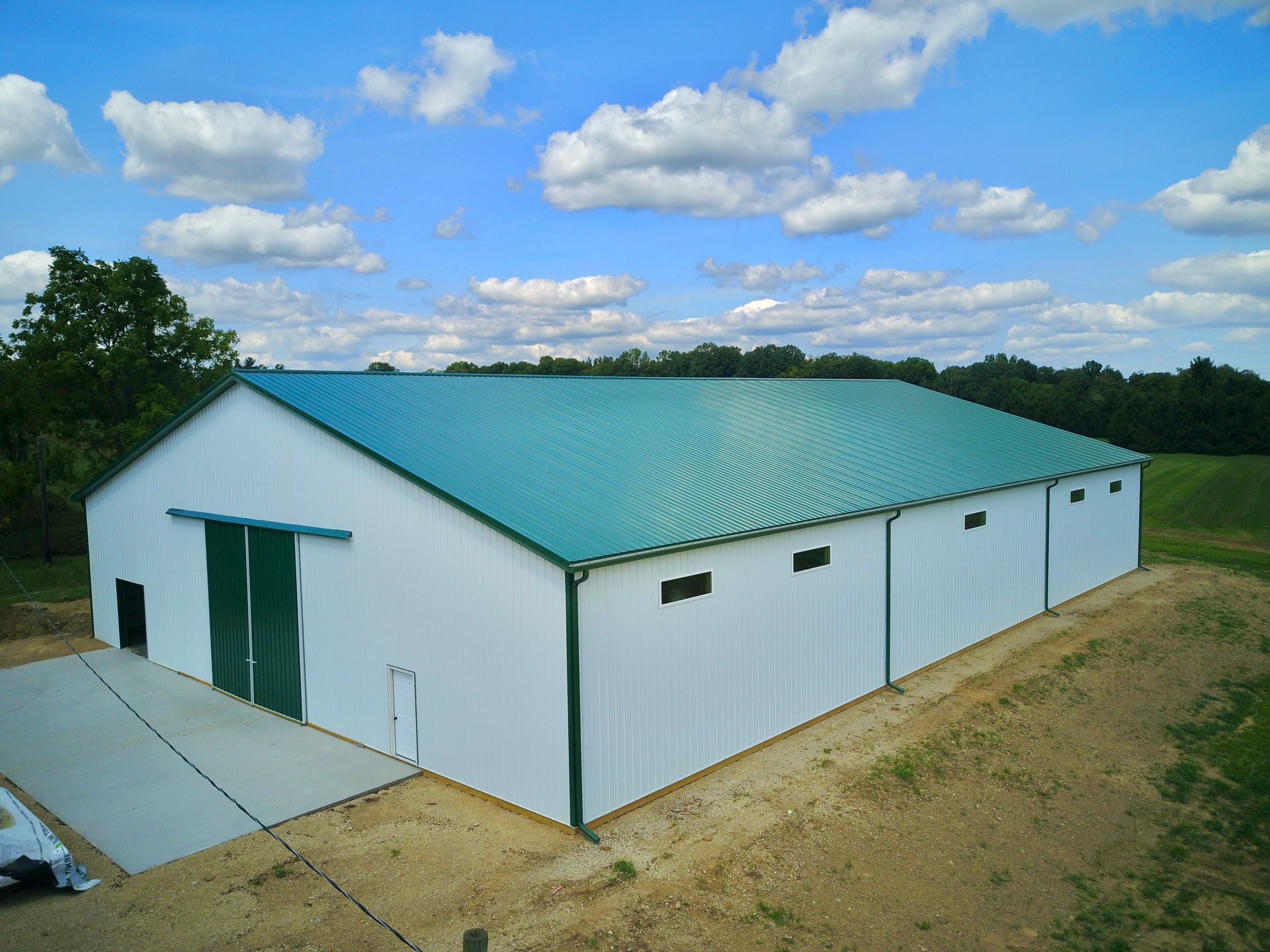 White metal barn with green trim and a teal roof, set on a grassy field under a blue sky with clouds.