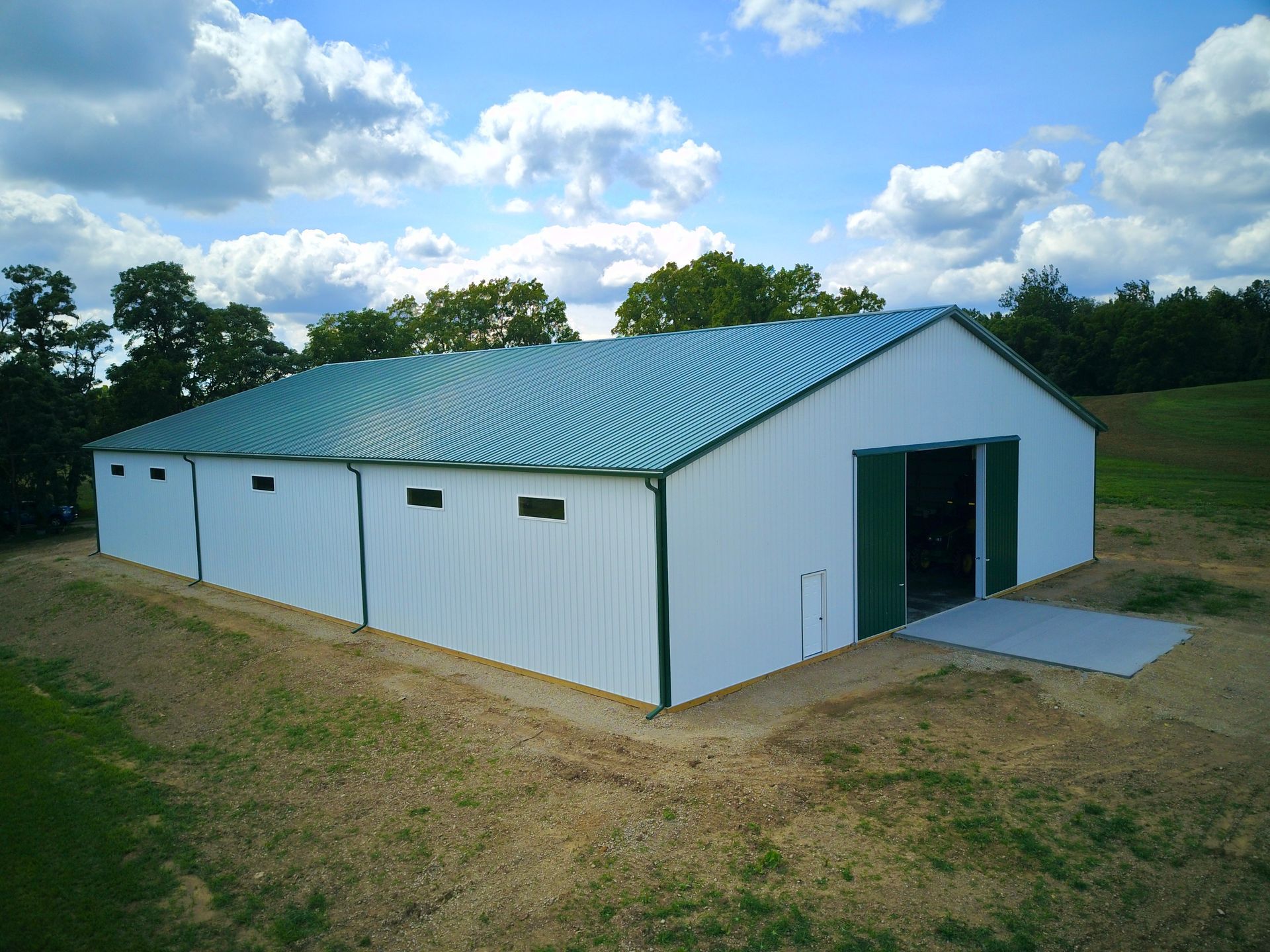 White and green metal agricultural building under a blue sky with trees. Doors are open.