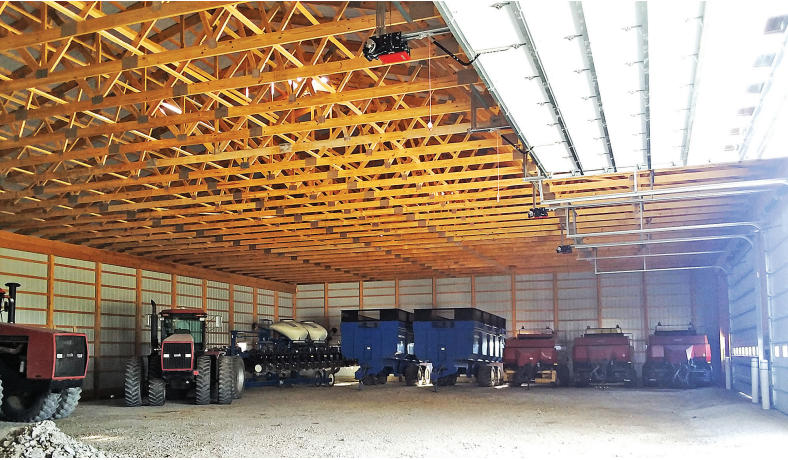 Inside of a large barn, tractors and farming equipment are parked. The building has a wooden truss roof and bright lights.
