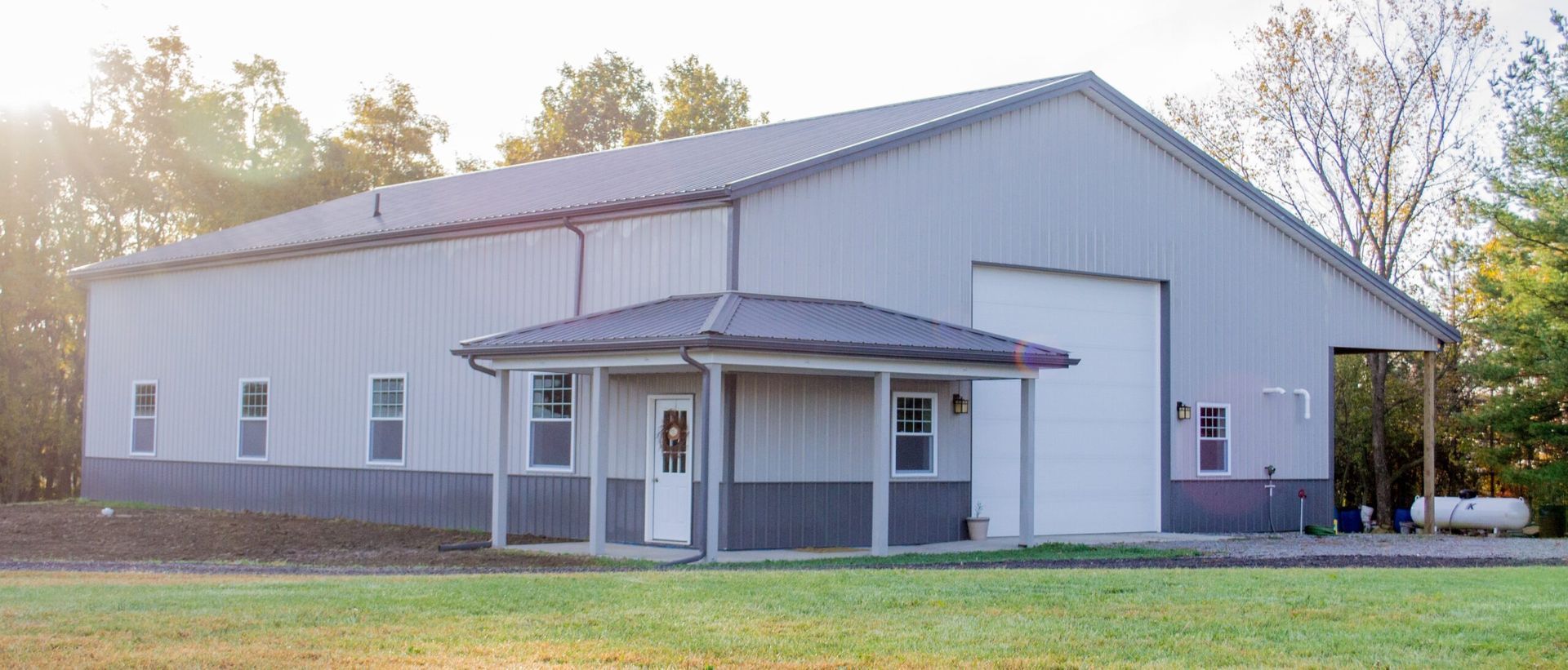 A large, gray metal building with a small covered porch and garage door. Trees and green grass surround the structure.