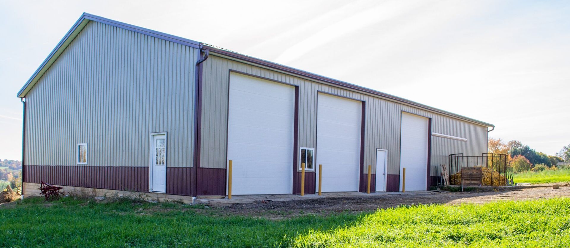 A long metal building with large garage doors sits in a grassy field. The building is light gray and has brown trim.