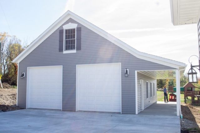 Two-car gray garage with a carport on a sunny day. A person stands near the carport entrance.