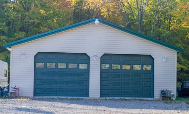 Two-car garage with teal doors and trim, beige siding, and a gravel driveway, set against a backdrop of fall foliage.