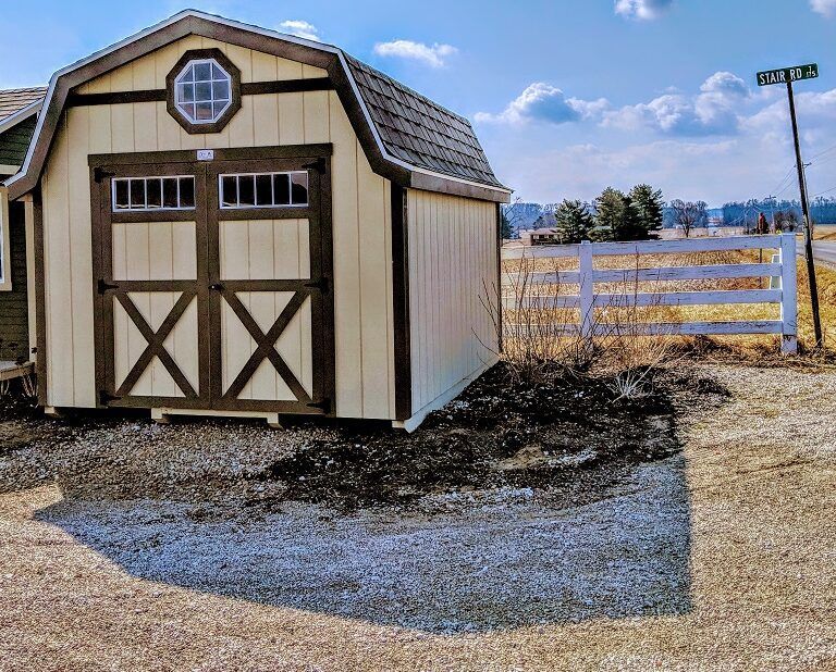 A beige barn-style shed with dark brown accents sits on a brown field, casting a large shadow.