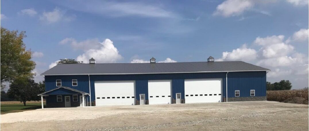 A large blue building with white garage doors sits under a partly cloudy sky. The building is in a rural setting.