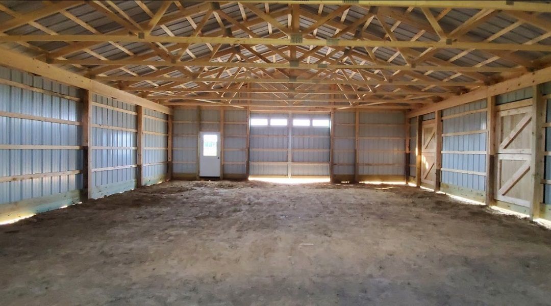 Interior view of a large, empty barn with wooden beams and metal siding. Sunlight streams through the open doors.