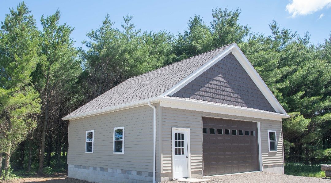 A gray-sided garage with a brown garage door, a white door, and two windows. A darker gray roof and trees in the background.