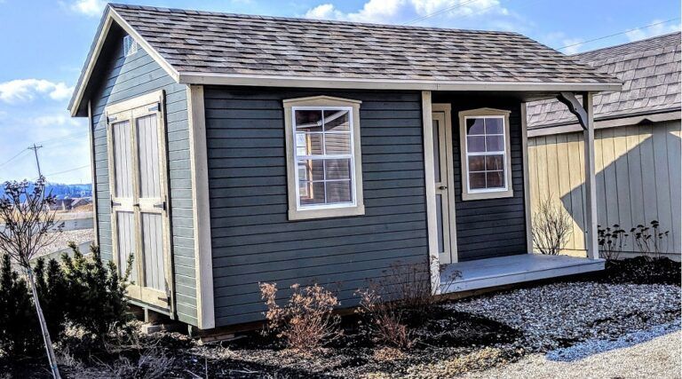 Small blue shed with white trim and a porch, set in a yard with a sunny sky in the background.