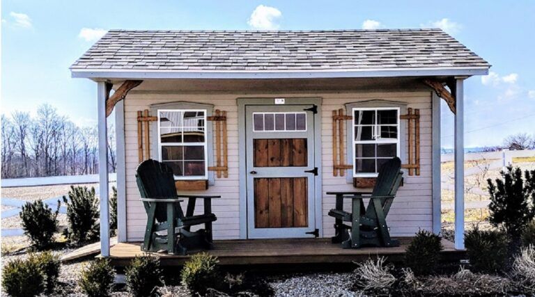Cozy, light pink shed with a wooden door and shutters. Two green Adirondack chairs sit on the porch. Snowy, rural setting.