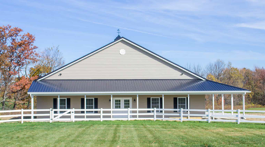 Tan building with dark roof and white porch, behind a white fence on a green lawn.