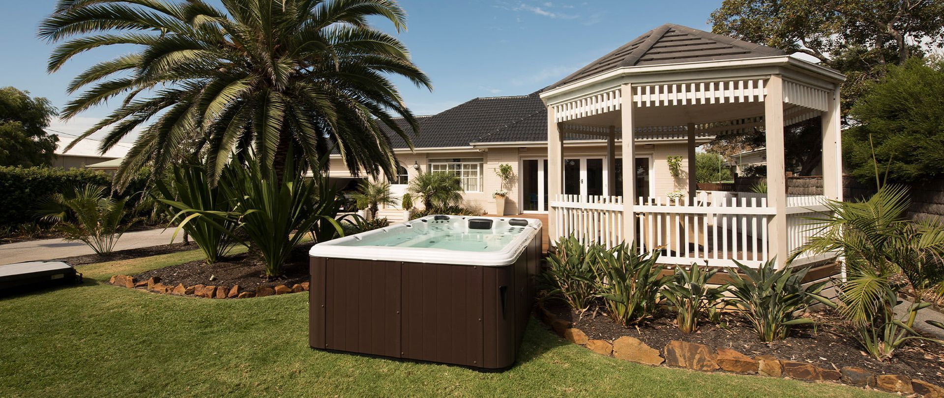 A brown hot tub in a backyard with a gazebo and palm tree.— Intouch Pools & Spas in Old Bar, NSW