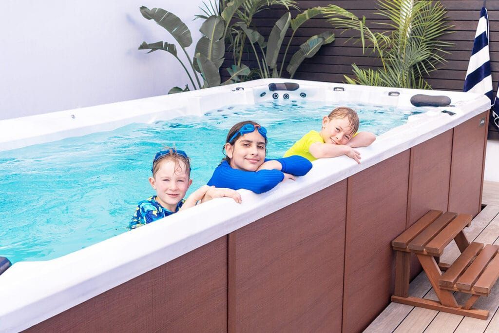 Three children smiling in a swim spa; brown paneling, wooden steps, plants in background. — Intouch Pools & Spas in Forster, NSW