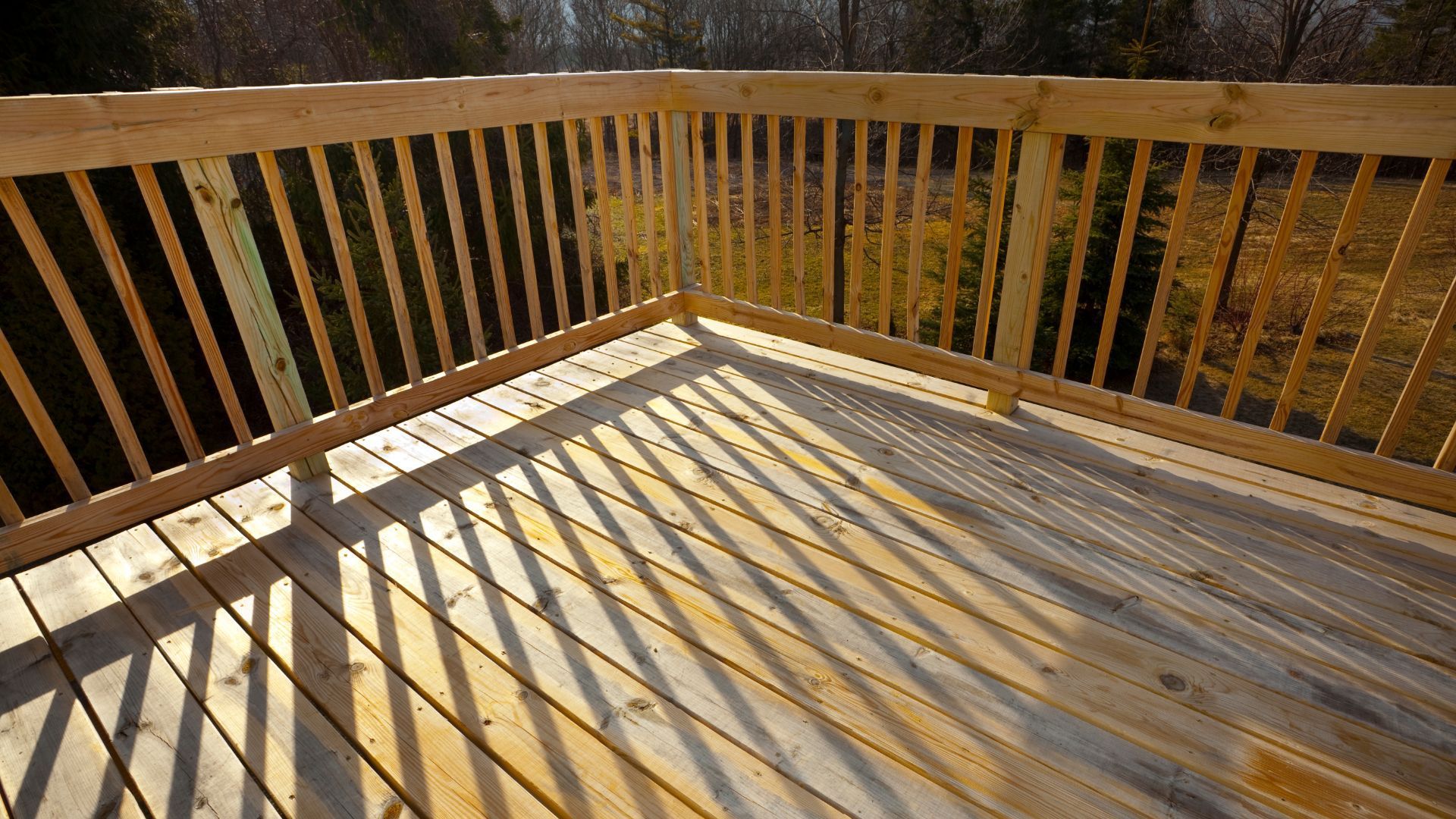 A sunlit wooden deck in Tempe, AZ captures the afternoon light, casting intricate shadows from the railings onto the deck floor. The natural tones of the wood decking blend harmoniously with the warm Arizona sun, creating a peaceful outdoor space. The surrounding landscape, visible through the railings, adds to the serene atmosphere, offering a private retreat for relaxation or outdoor activities. This deck exemplifies the charm of outdoor living spaces in Tempe.