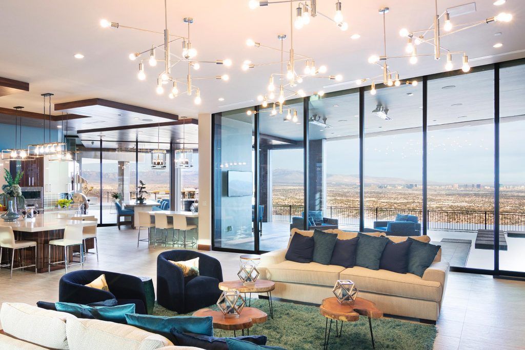 Spacious modern living room with large windows, beige sofa, teal accents, and numerous hanging lights overlooking a cityscape.