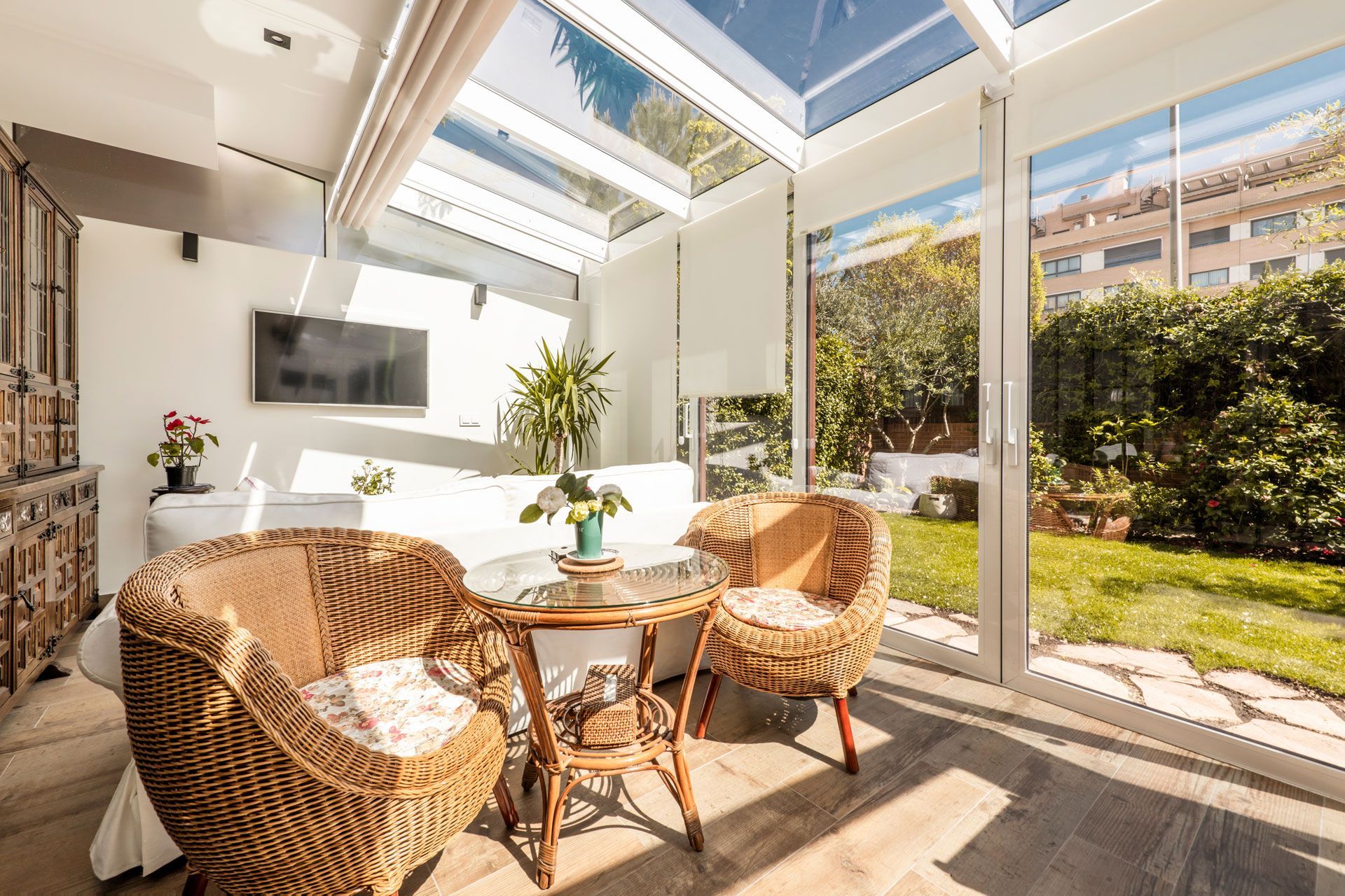 Sunroom with wicker chairs, small table, and glass roof overlooking a garden.