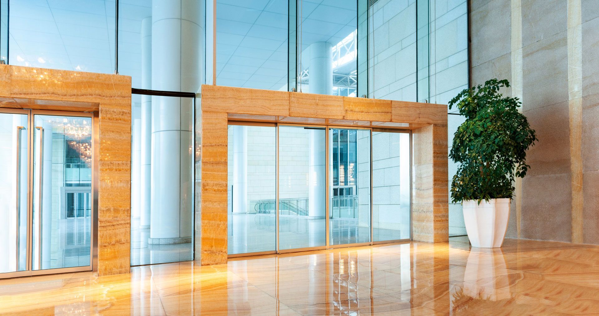 Sliding glass doors framed by beige stone in a modern building lobby, with a potted plant to the right.