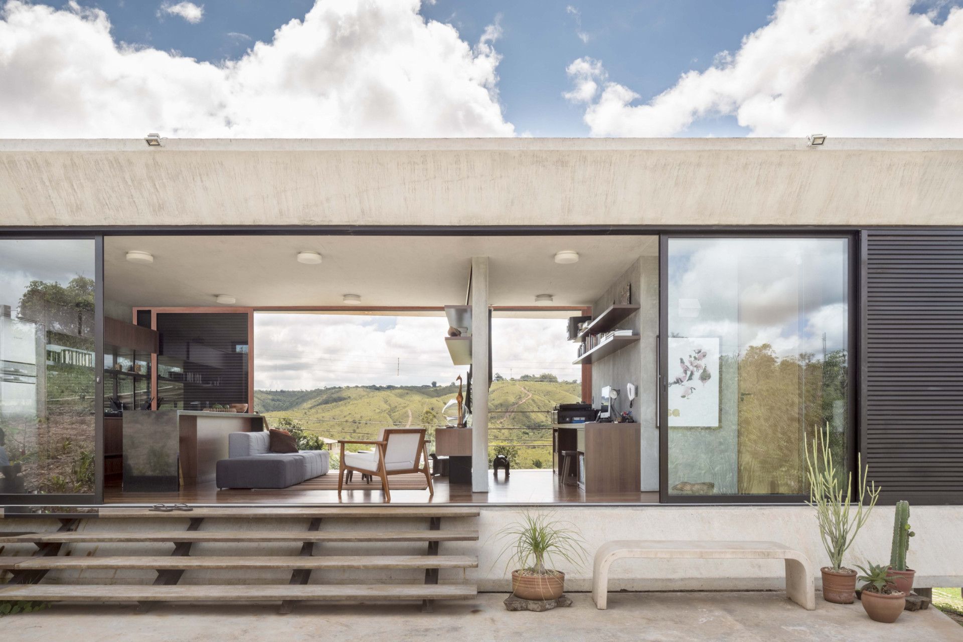 Modern house with large windows, open to the outside. Concrete steps lead to living area with hillside view.