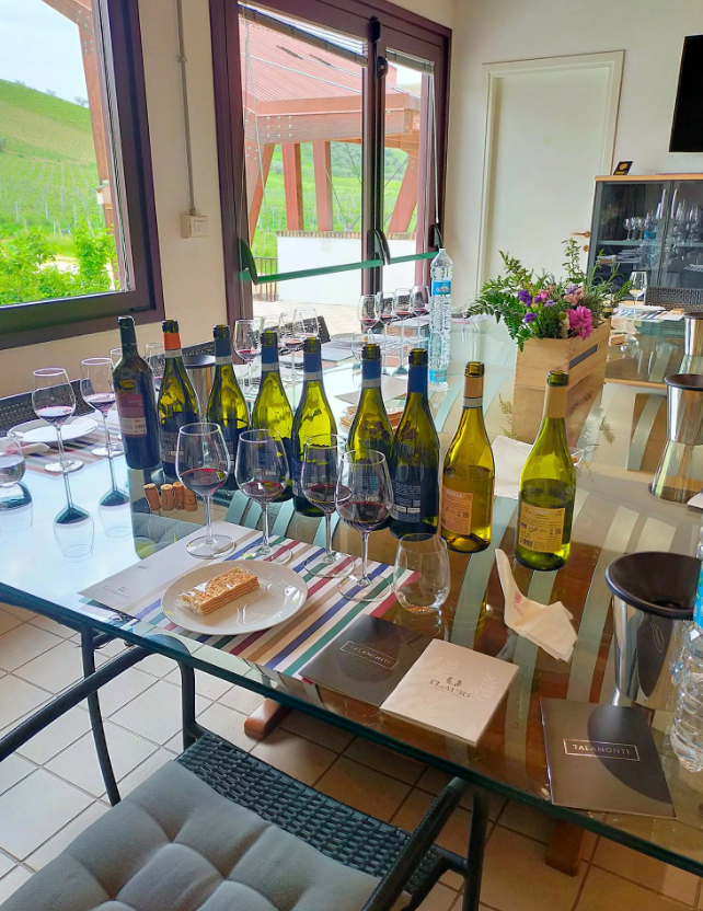 Un tavolo per la degustazione di vini, con diverse bottiglie, bicchieri pieni e piccoli stuzzichini, in una stanza con ampie finestre.