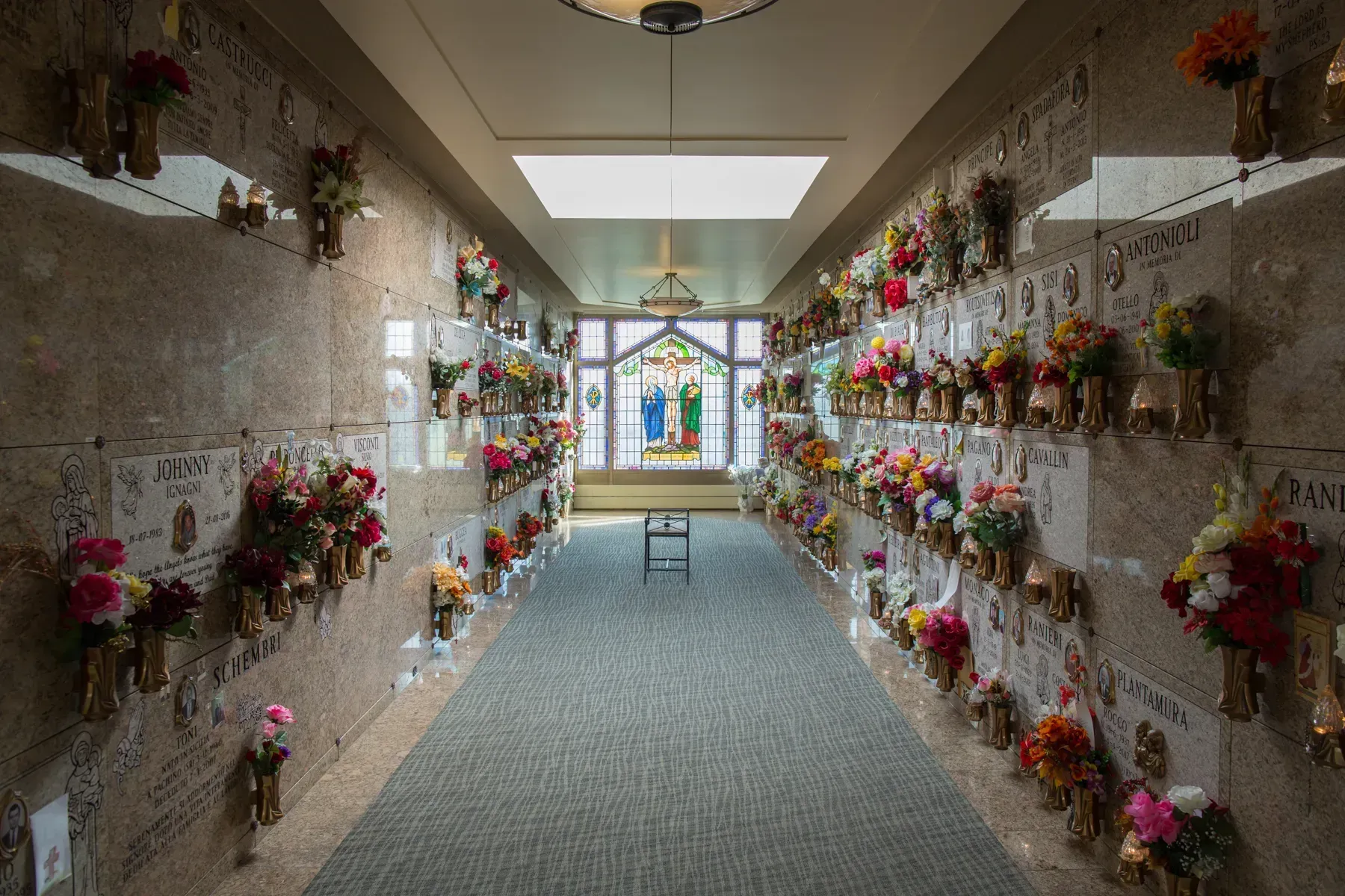 Narrow mausoleum hallway lined with floral arrangements on crypt walls, leading to a bright cross at the end.