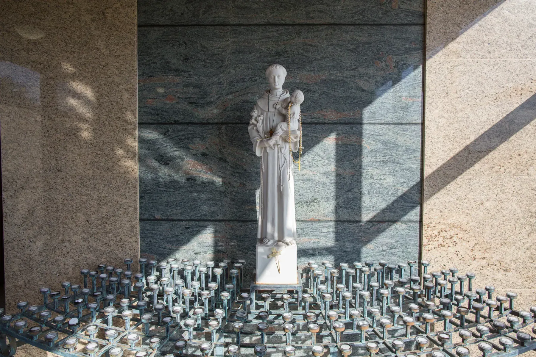 White statue in a memorial niche, surrounded by many lit candles on a stone floor.