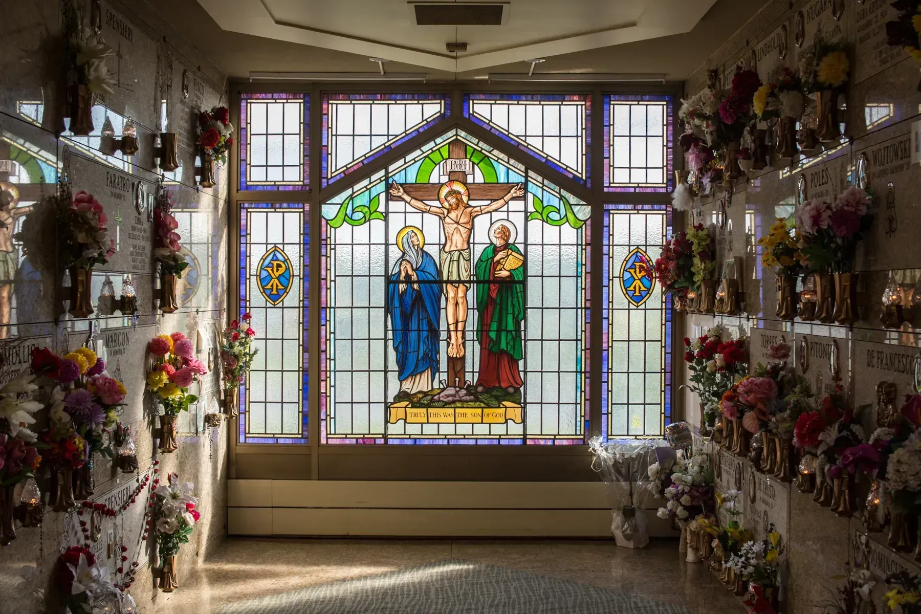 Stained-glass memorial display with flowers lining both walls in a chapel-like hallway.