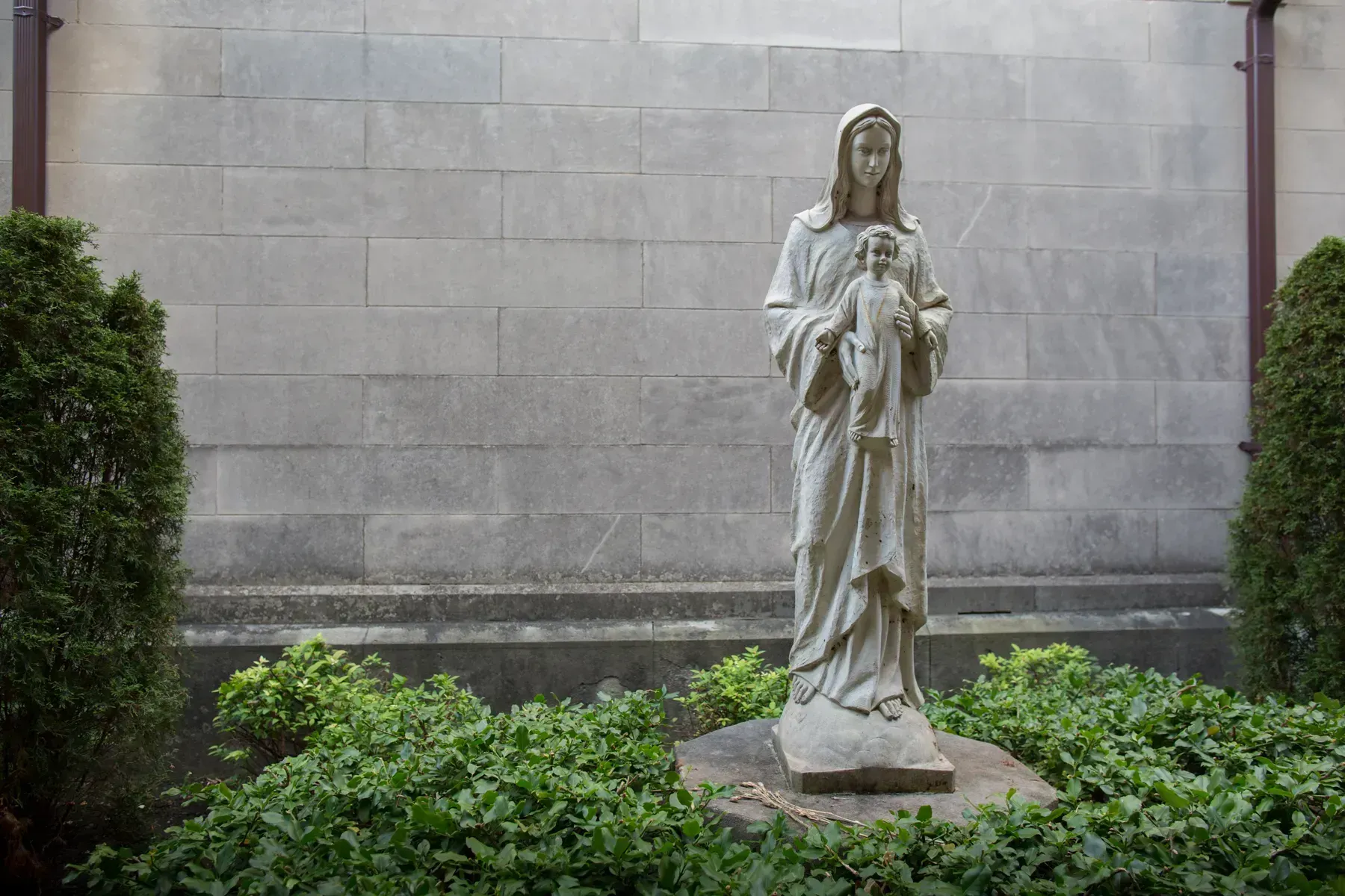 Stone statue of a robed figure holding a child, set in a garden against a gray wall.