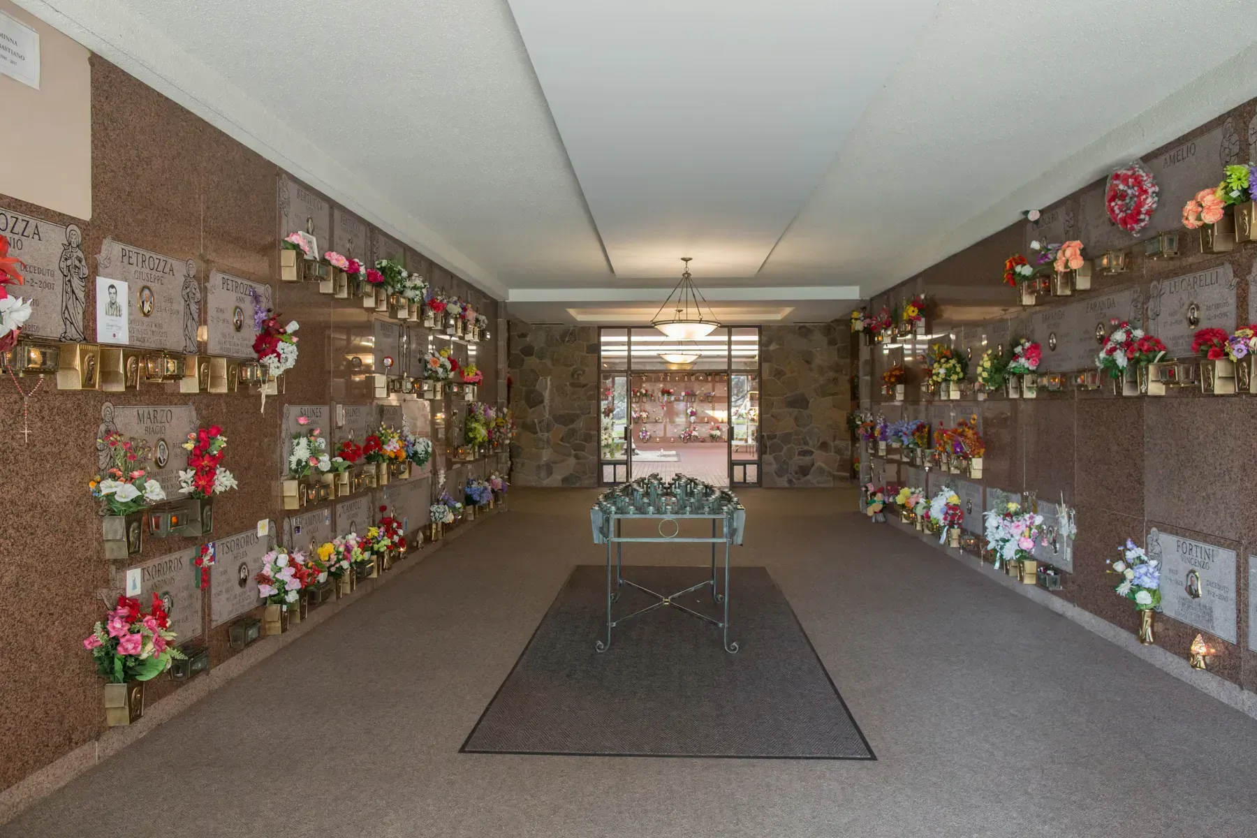 Flower-lined hallway with rows of memorial plaques and a central table on a carpeted floor