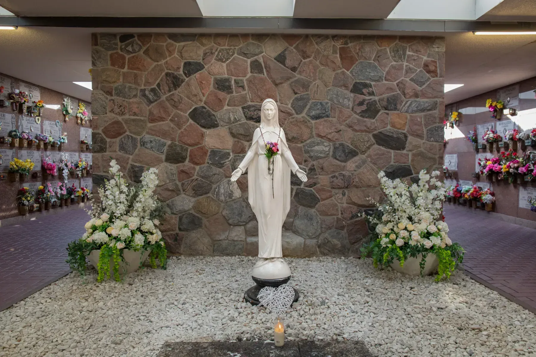 Statue of the Virgin Mary in a floral memorial shrine with stone wall backdrop