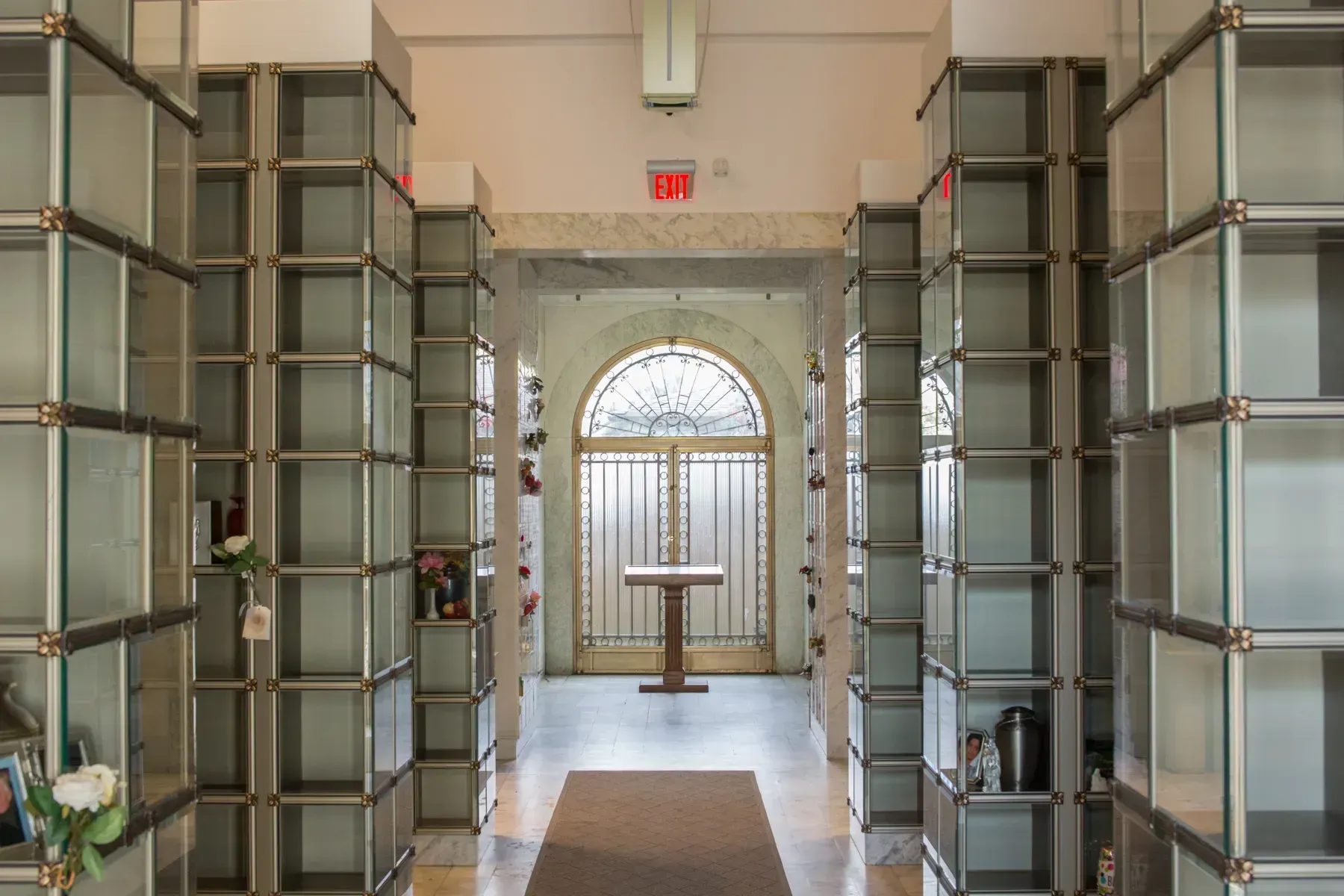 Interior hallway with rows of glass-front cremation niches and a central memorial table near a stained-glass window.