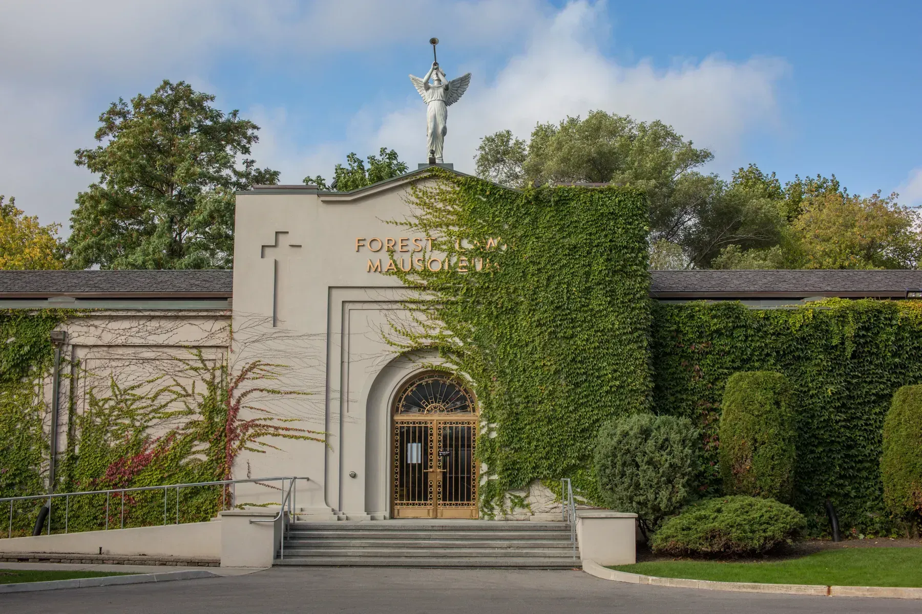 Stone building entrance with arched doorway, cross, and ivy-covered wall amid trees and shrubs