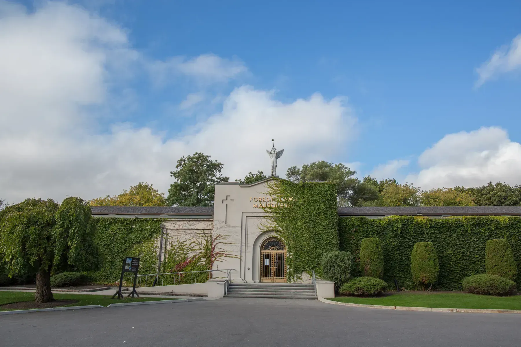 Stone entrance gate with ivy-covered hedges under a blue sky with clouds