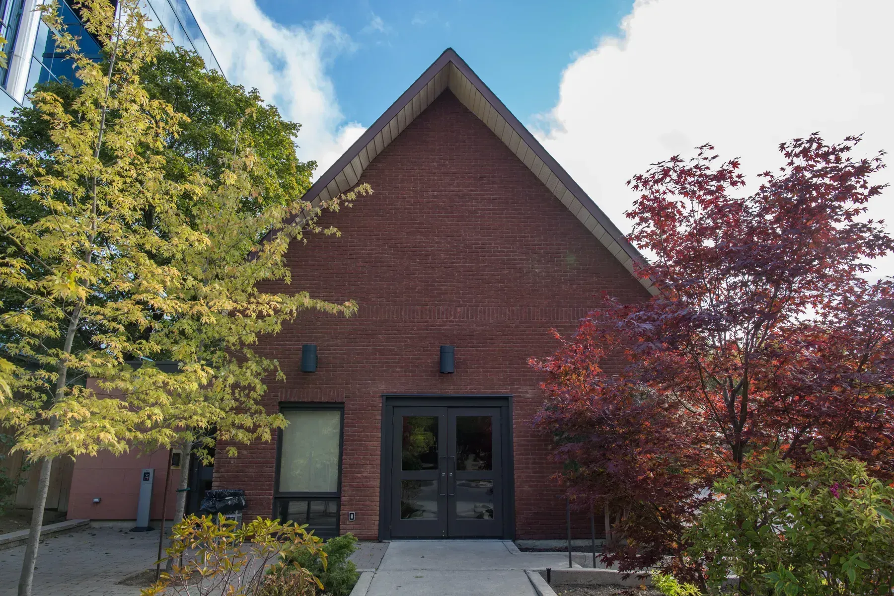 Brick building entrance with dark double doors, framed by green and red trees.