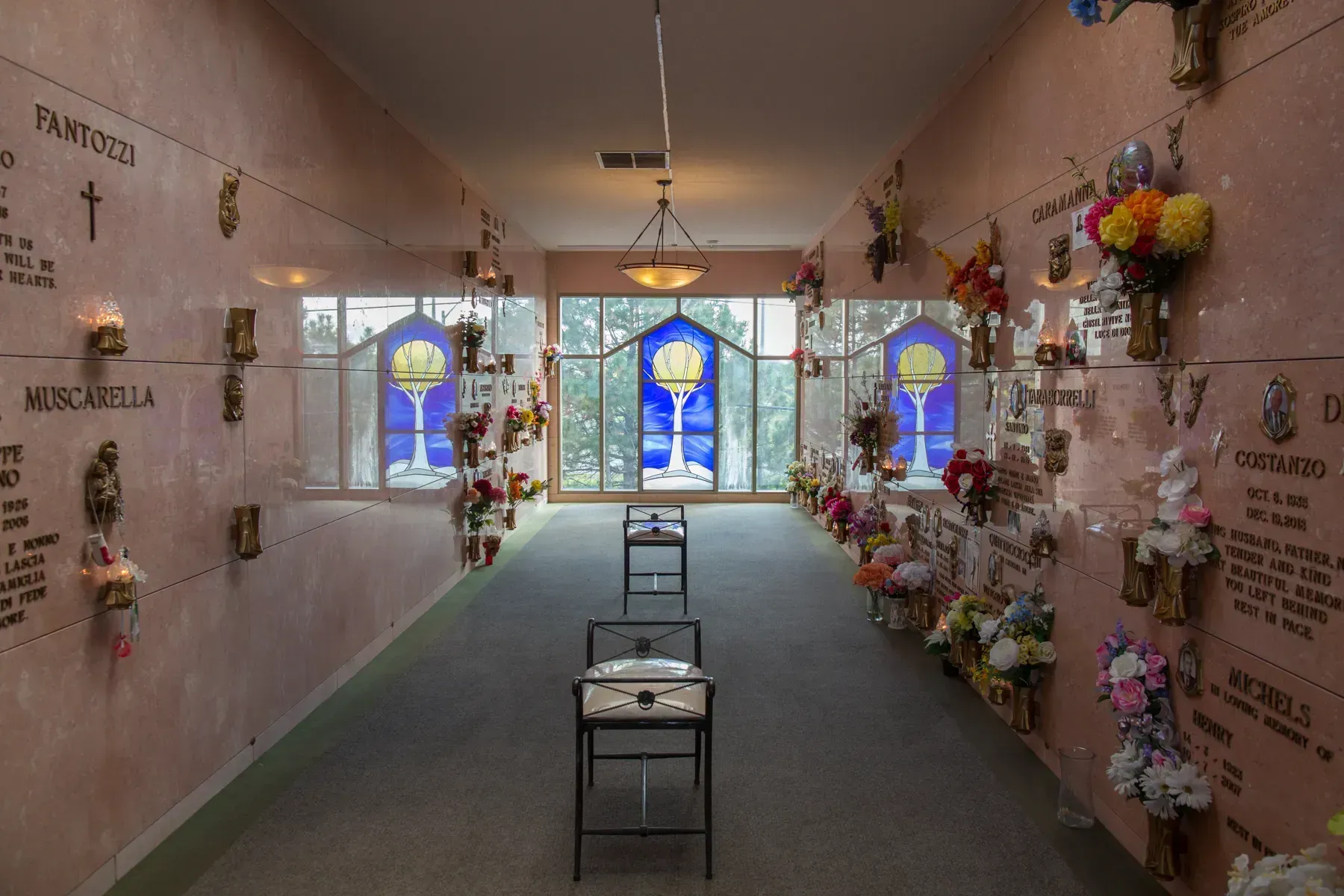 Narrow memorial hallway with colorful flower tributes, plaques, and stained-glass windows at the end