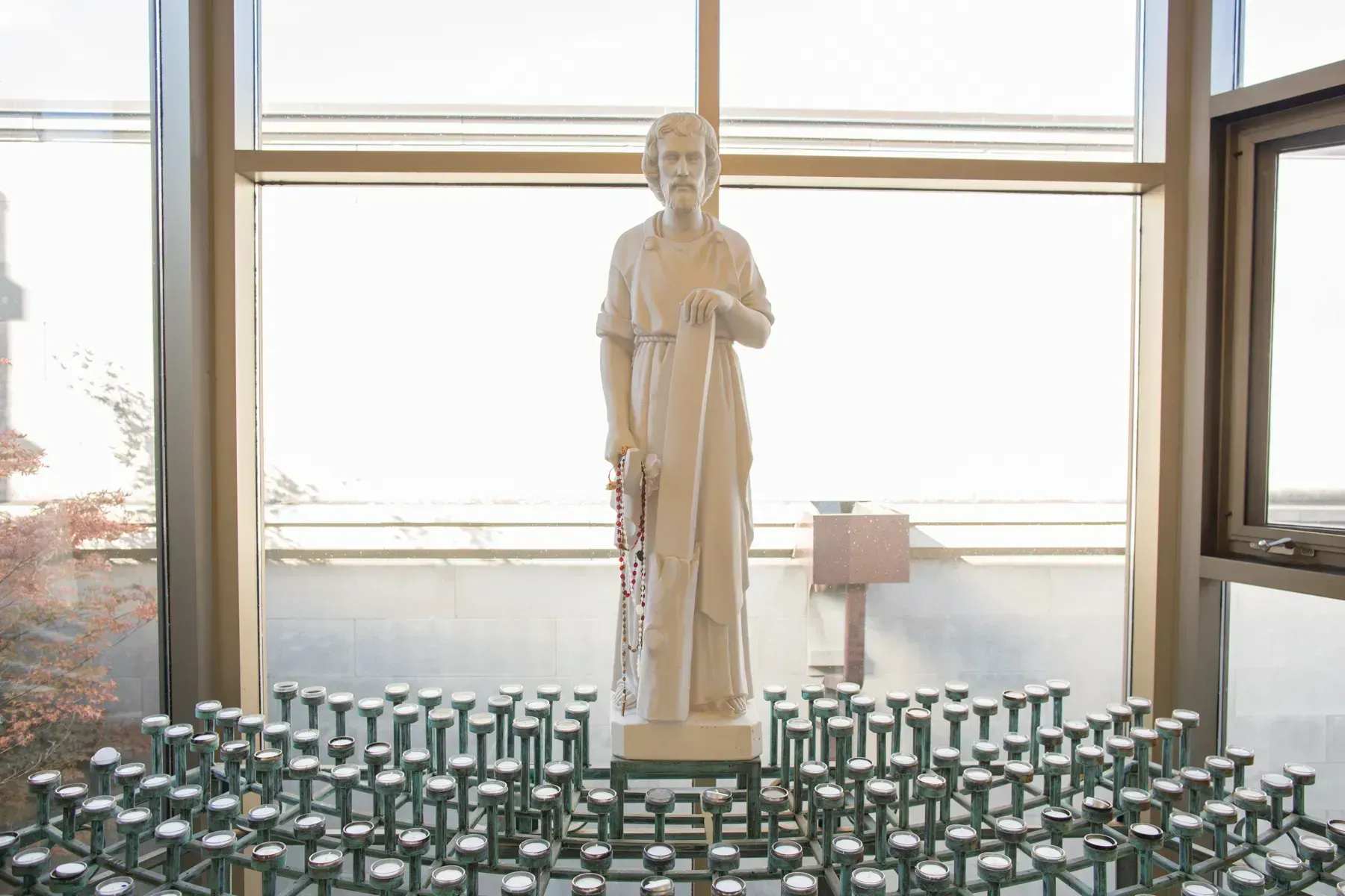 White statue on a pedestal before large windows, surrounded by rows of green votive candles