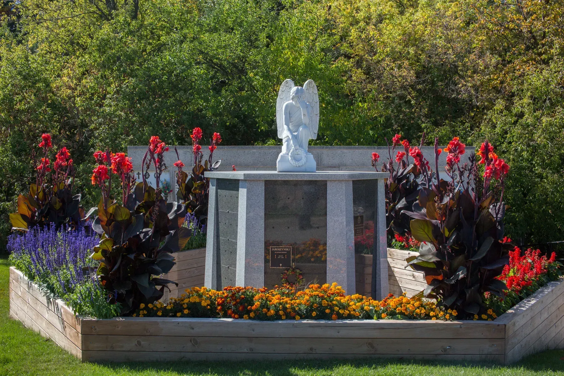 Memorial garden with a white statue, stone plaque, and colorful flowers bordered by red and purple plants