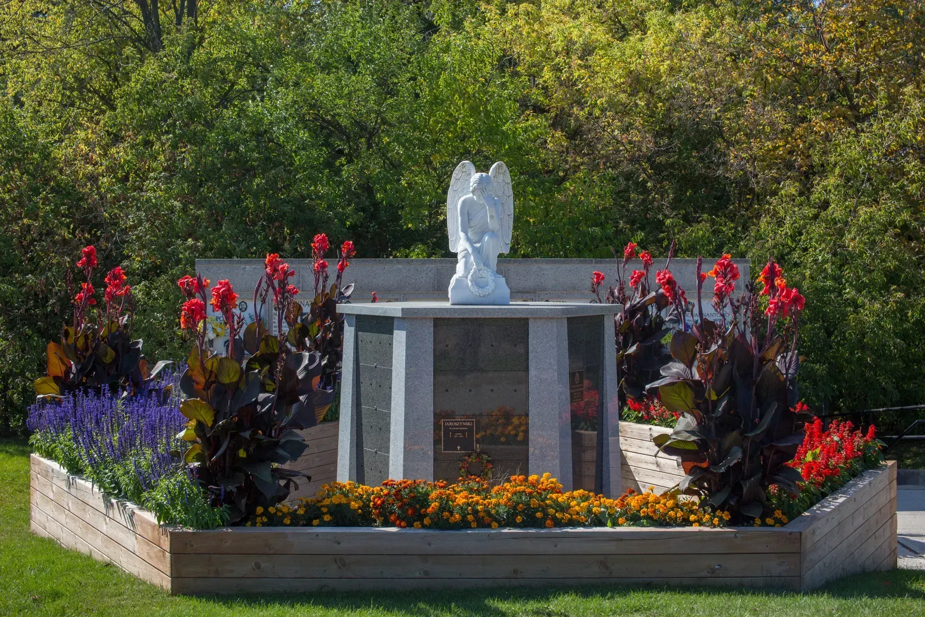 Memorial flower bed with a stone pedestal and white angel statue surrounded by red, purple, and orange flowers