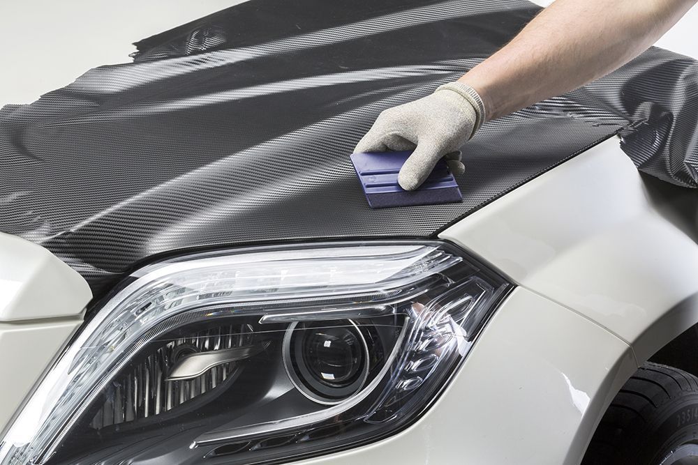Person applying a carbon fiber wrap to a car hood using a squeegee.