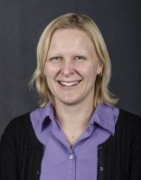 Woman smiling, wearing a purple shirt and black cardigan, against a dark background.