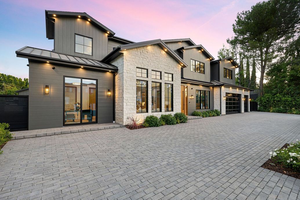 Modern two-story house with gray and brick facade, cobblestone driveway, and evening sky.
