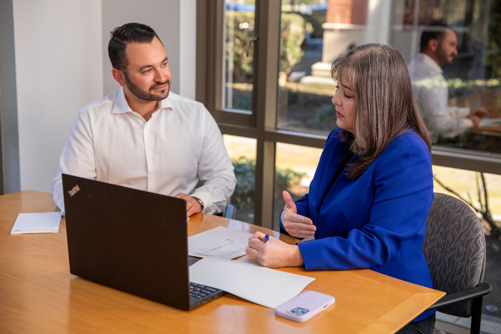 Two people in a meeting, one speaking with a laptop and documents on the table.