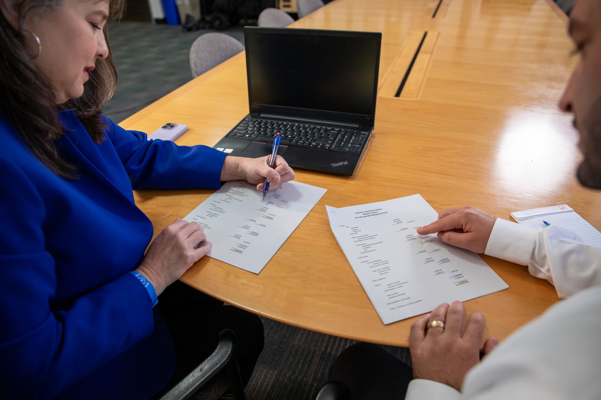 Two people working on papers at a table with a laptop in a classroom or office setting