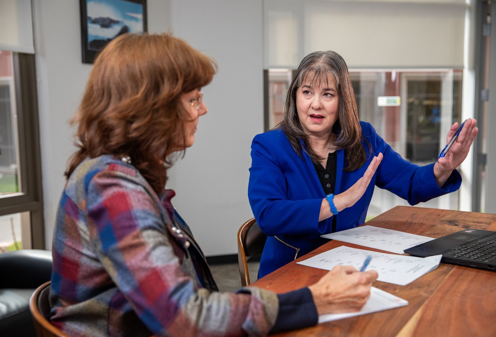Two women discussing paperwork at a table in a bright office, one gesturing while speaking.