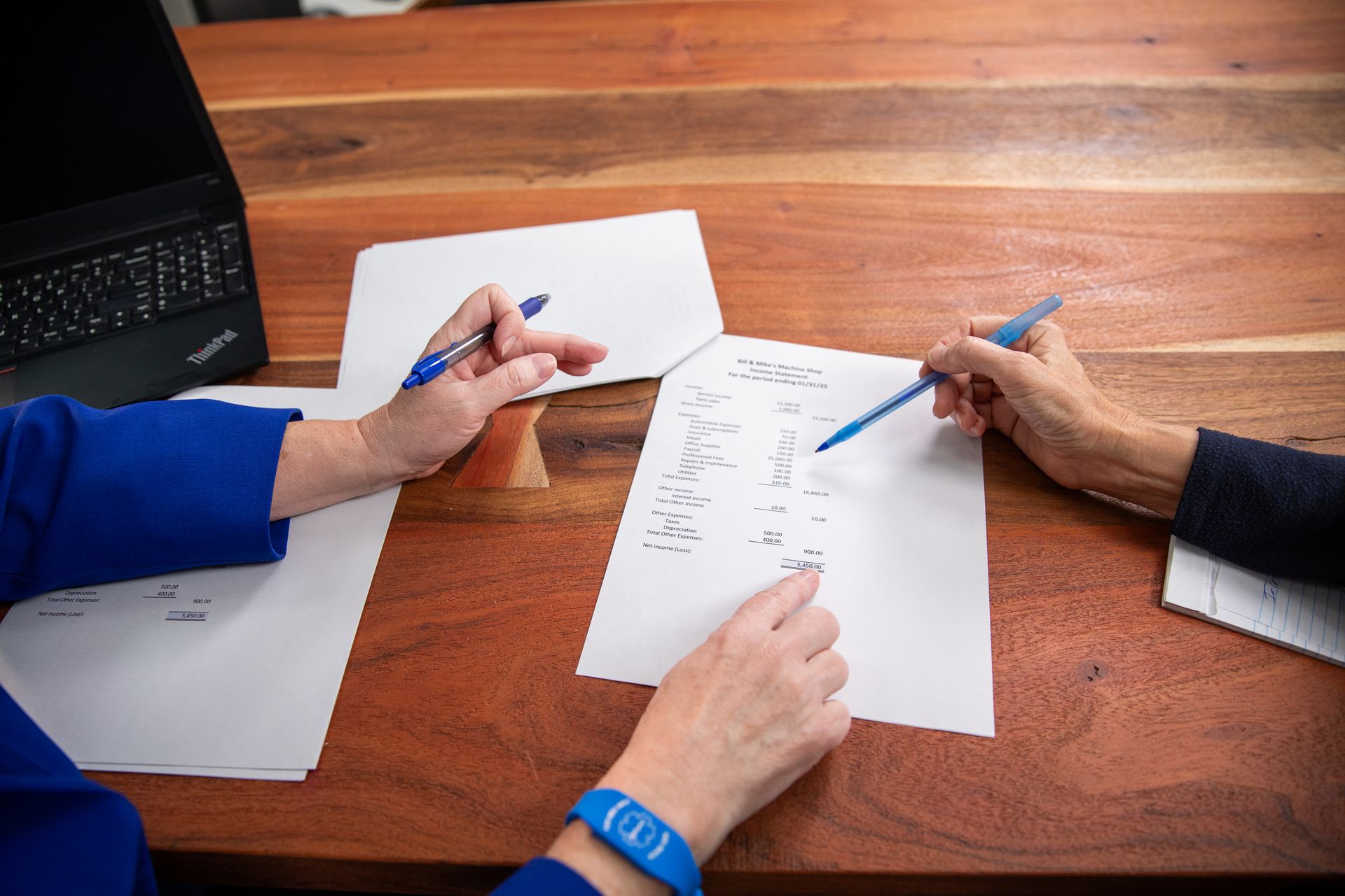 Two people with pens reviewing papers at a wooden table. One points to the paper while the other holds a pen.