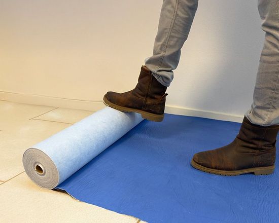 Een persoon in laarzen stapt op een uitgerolde blauwe mat. De mat ligt uitgerold op een licht betegelde vloer.