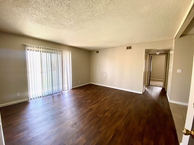 Empty living room with dark wood-look flooring, sliding glass door, and neutral-colored walls.