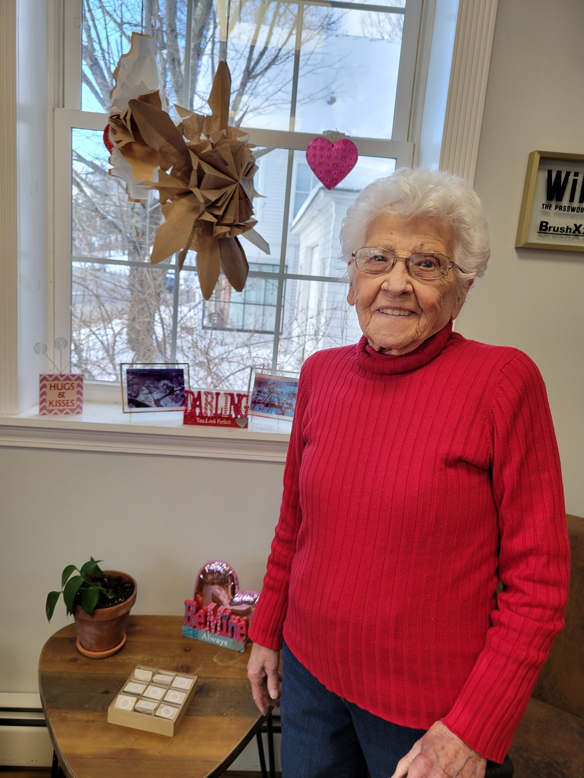 An elderly woman in a red sweater is standing in front of a window.