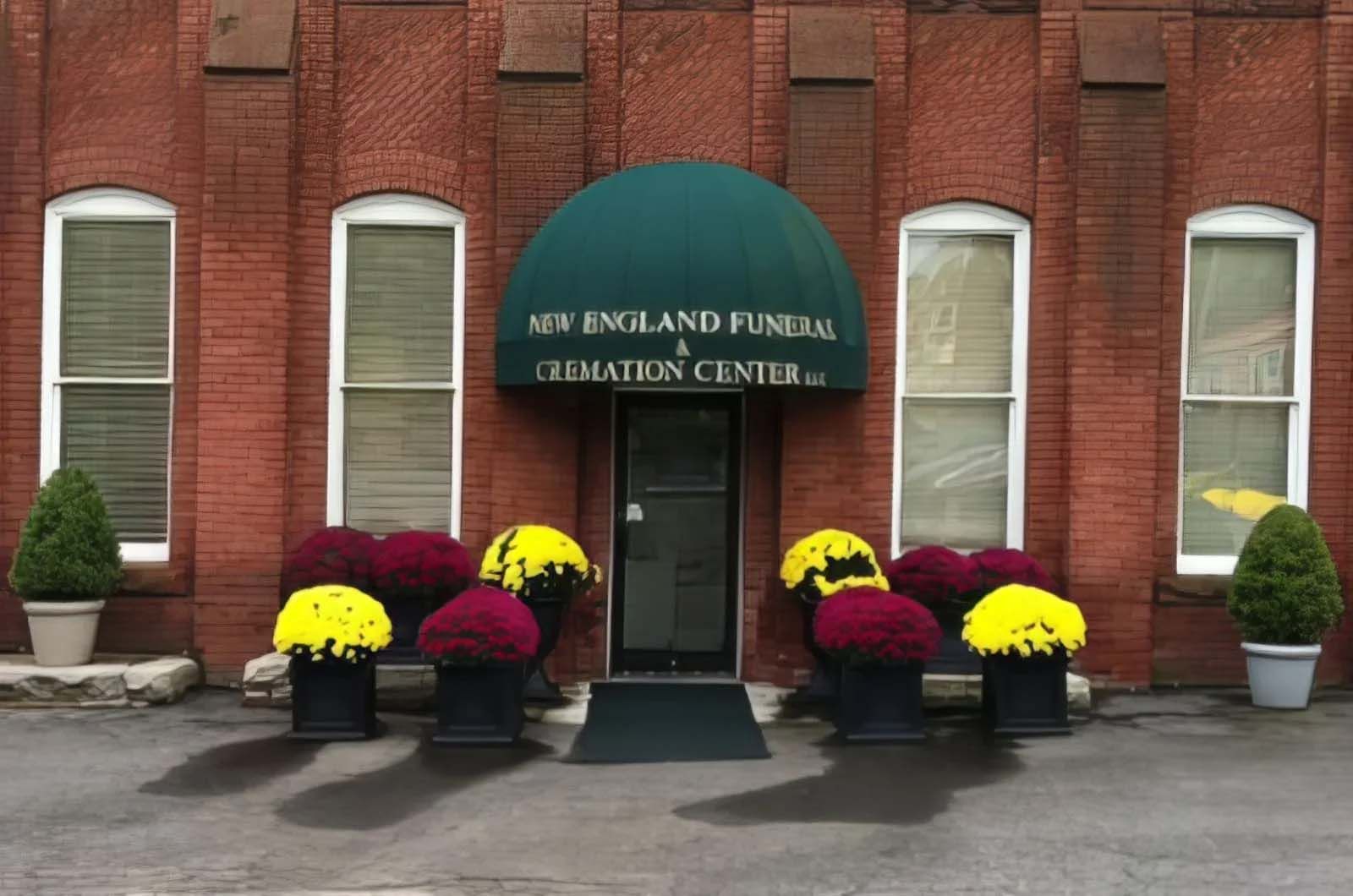 New England Funeral & Cremation Center entrance with a green awning, brick facade, and colorful mums in planters.